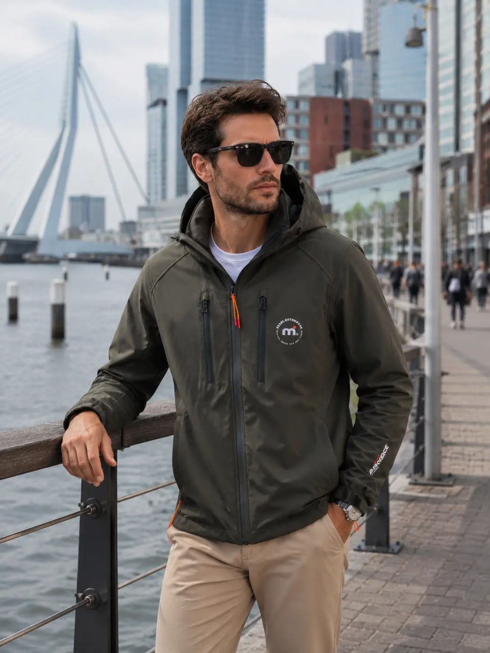 Man wearing a green jacket with a logo, sunglasses, and beige pants by a waterfront with a bridge and buildings in the background.