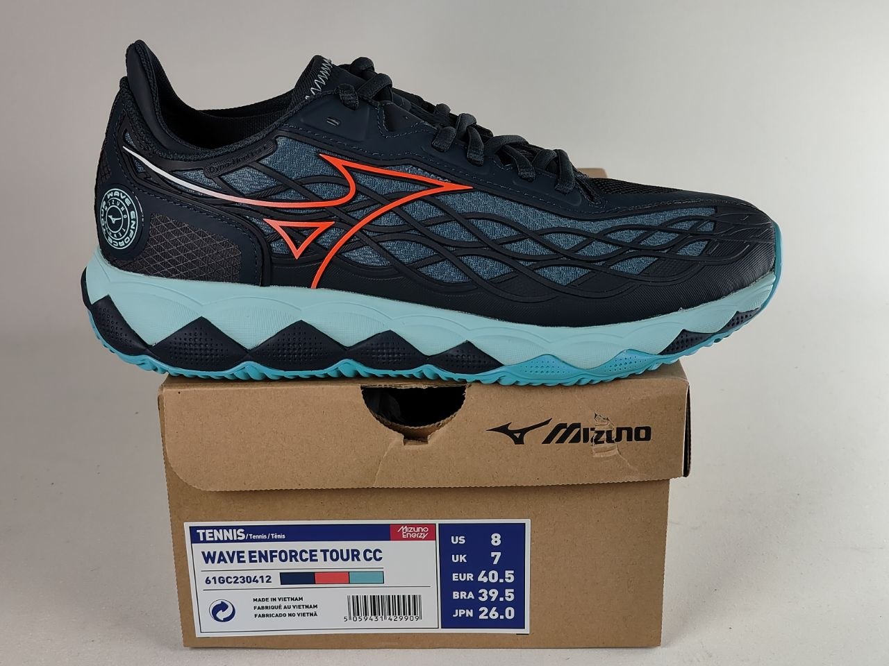 A Mizuno tennis shoe in navy blue with orange and light blue accents, displayed on top of its original cardboard box with a size label visible.