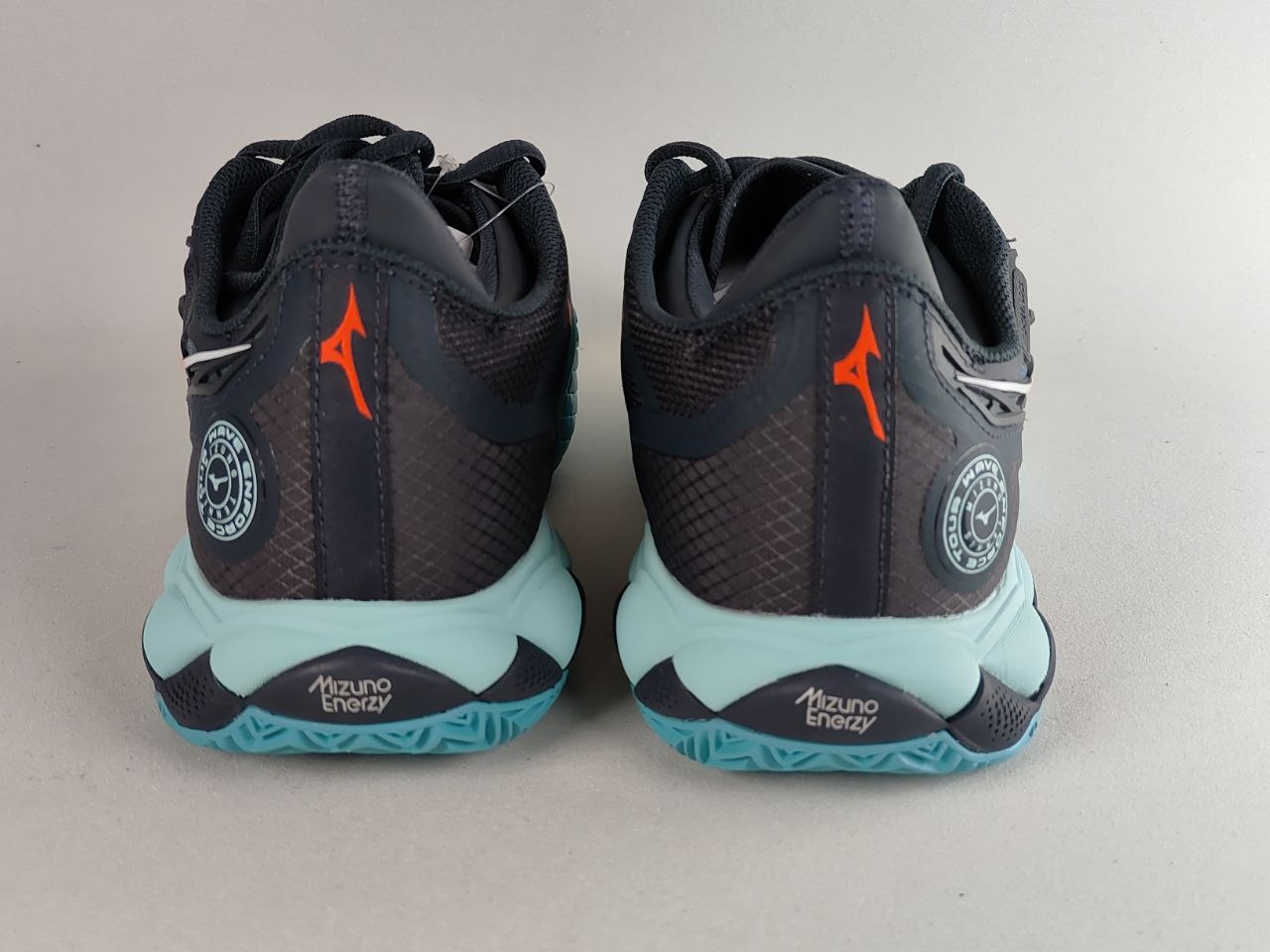 A pair of Mizuno athletic shoes shown from the back, featuring a dark navy and black mesh upper with bright orange logo accents, a light blue midsole, and a teal outsole.