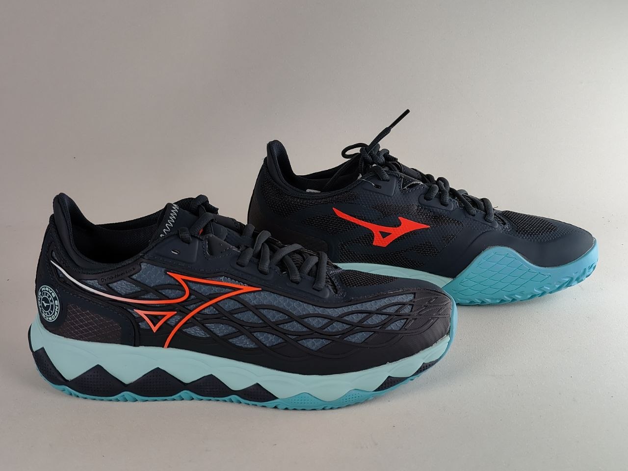 A pair of Mizuno athletic shoes in black and dark gray with bright orange logos and light blue soles, displayed against a neutral beige background.