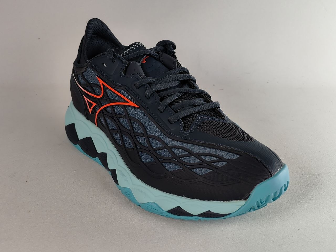 A Mizuno running shoe in black with a bright orange logo, a black and gray wavy mesh upper, and a light blue and black wavy sole, shown on a neutral light gray background.