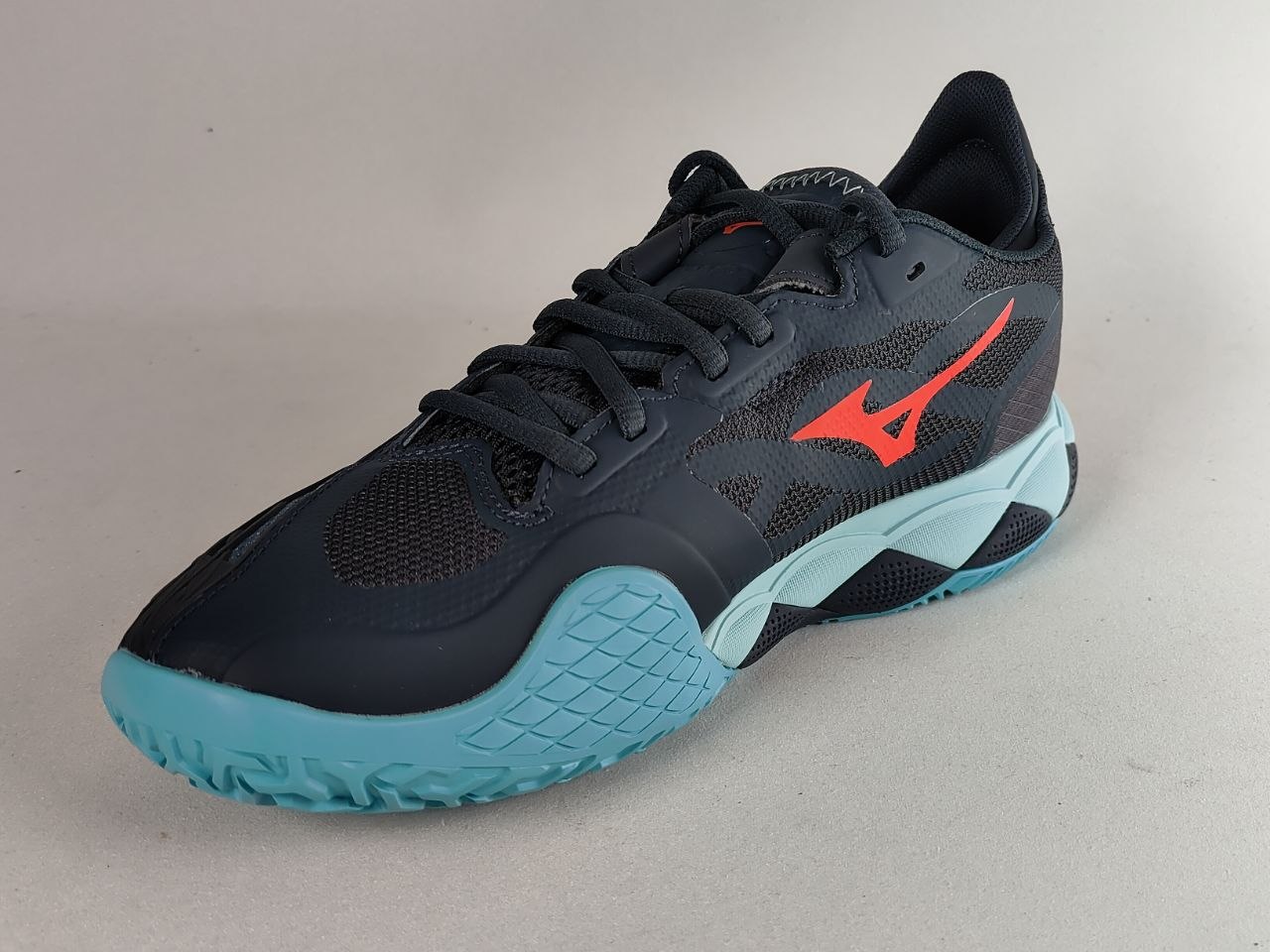 A Mizuno athletic shoe in black with a bright orange logo and a light blue sole, photographed on a neutral light gray background.