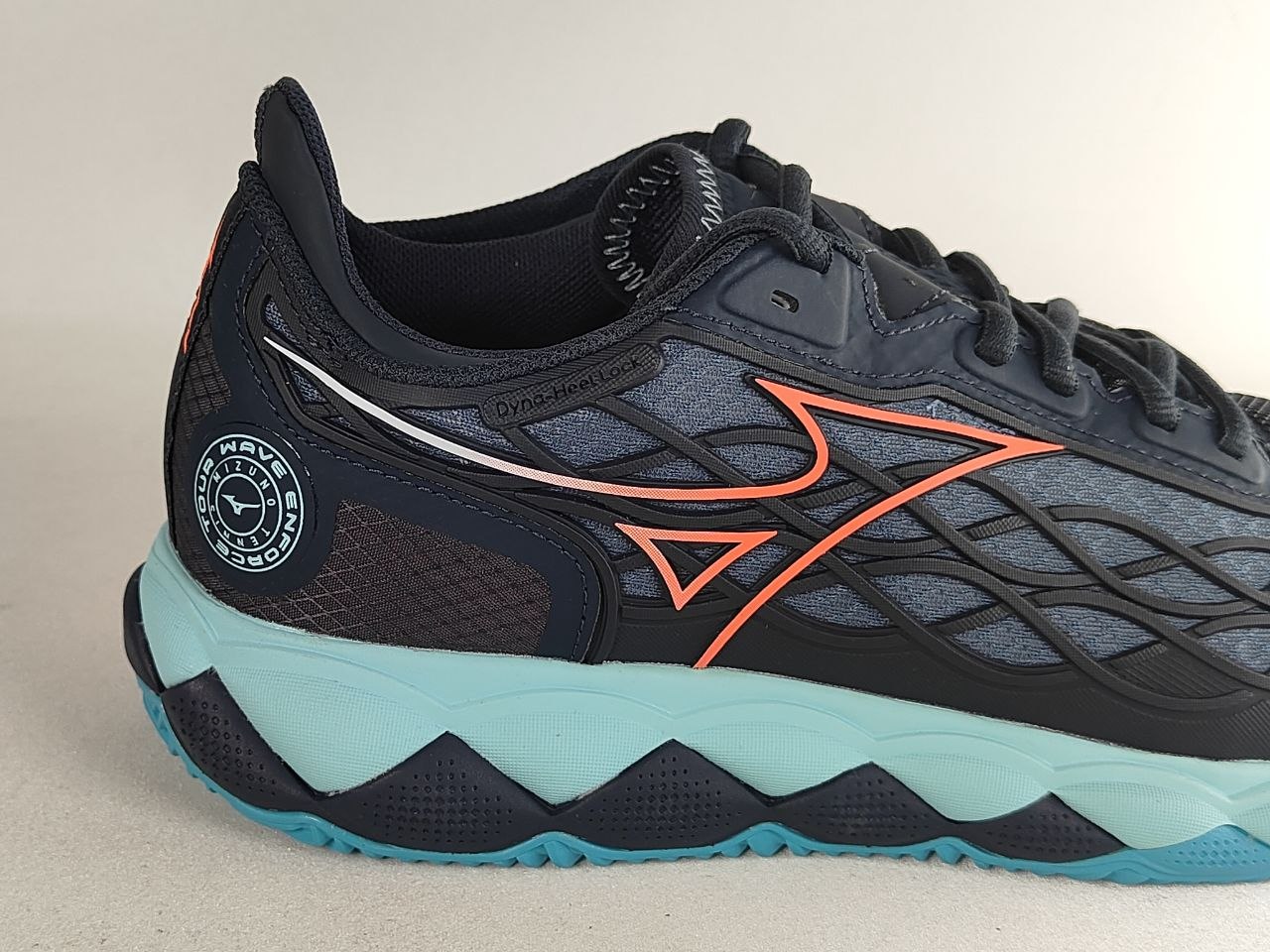 A close-up side view of a Mizuno running shoe in dark navy and gray with bright orange logo accents and a light blue and navy wavy midsole.