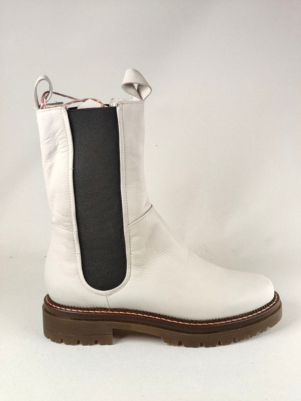 A white leather Chelsea-style ankle boot with a black elastic side panel and a brown lug sole, photographed against a light gray studio background.