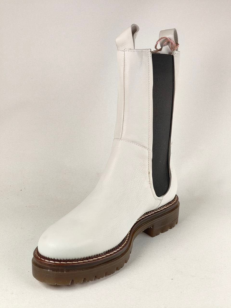A white leather ankle boot with a black elastic side panel and a brown lug sole, photographed against a light gray studio background.