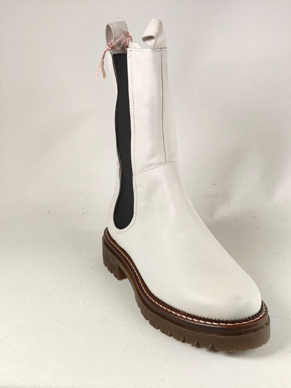 A white leather Chelsea boot with a thick brown lug sole, featuring a black elastic side panel and a small pink ribbon detail at the pull tab, photographed against a light gray background.