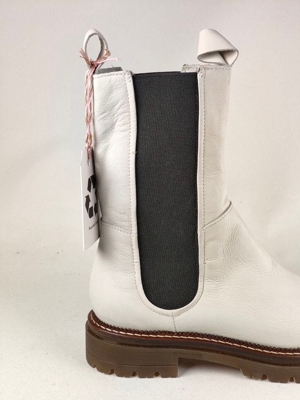 A white leather Chelsea boot with a black elastic side panel and a brown lug sole, shown on a light gray background with a hangtag attached.