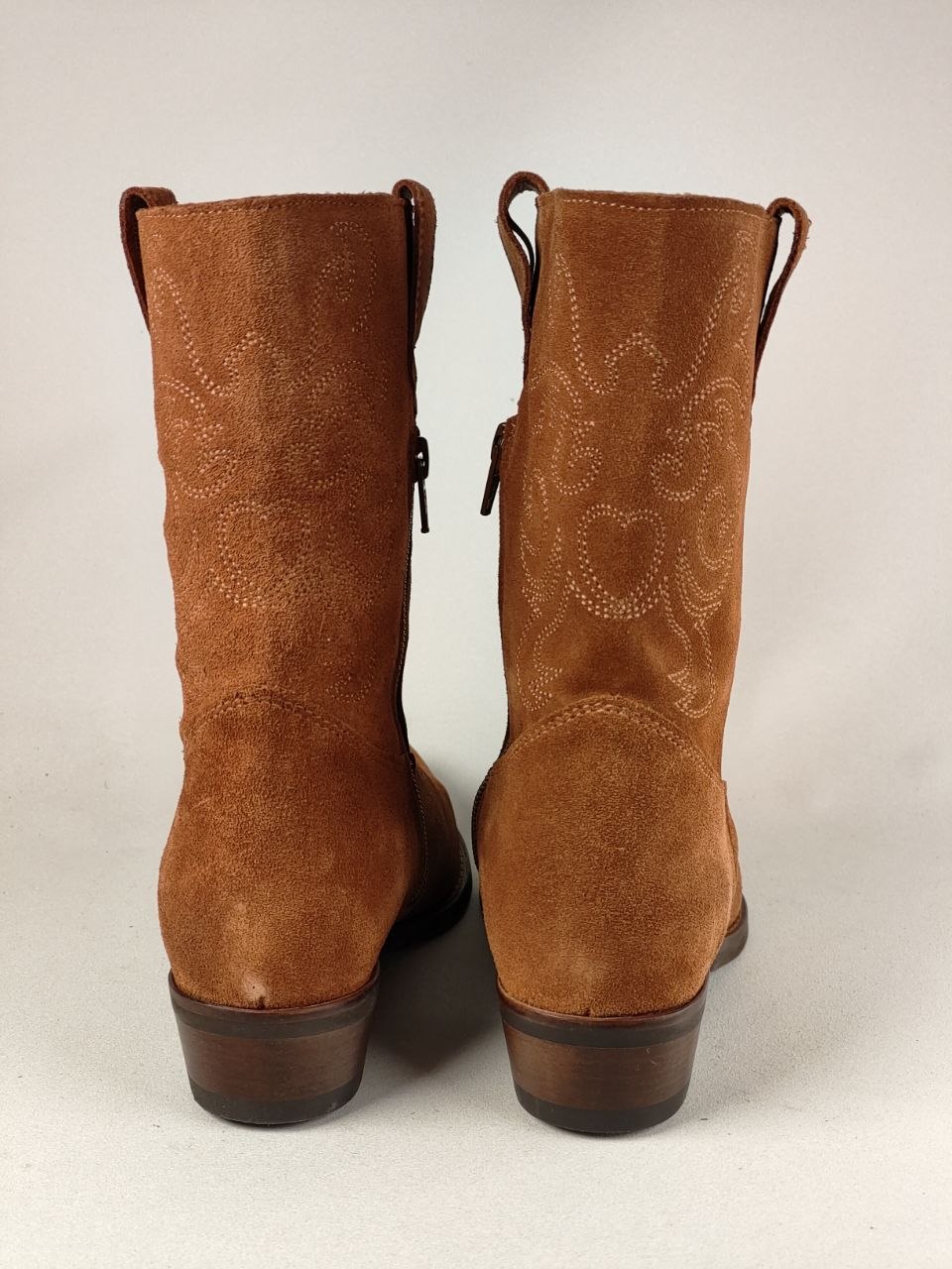 A pair of brown suede western boots shown from the back, featuring decorative cutout embroidery and stacked dark brown heels, set against a plain light background.