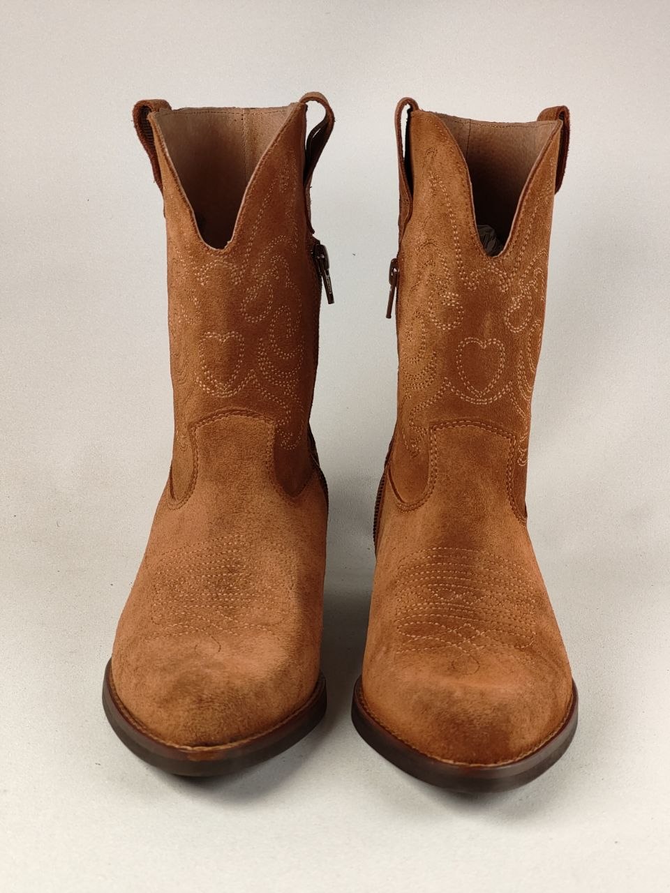 A pair of tan suede western ankle boots with decorative stitching on the shafts, shown from the front against a light gray background.