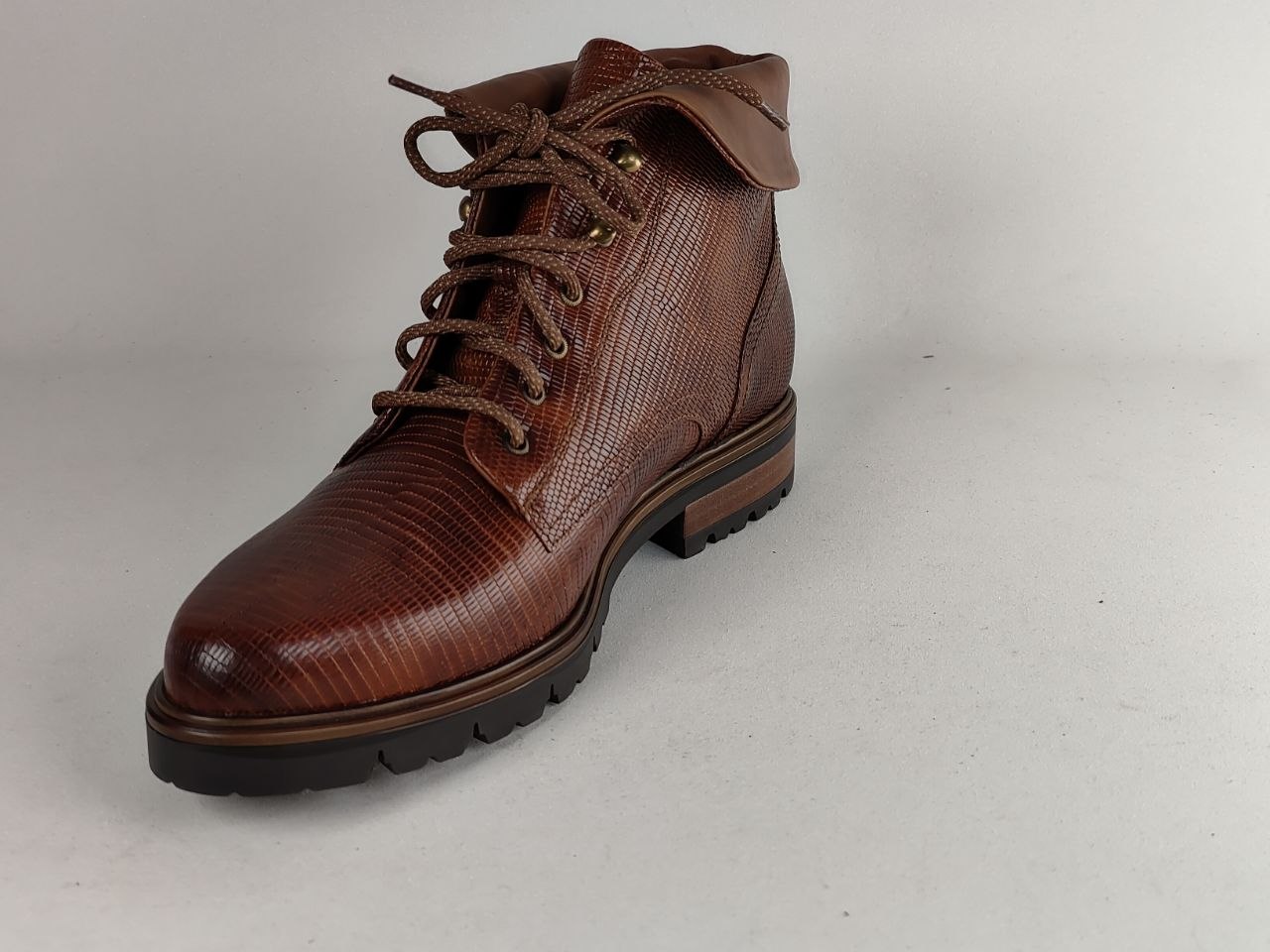 A brown textured leather lace-up ankle boot with a rugged black sole, photographed on a neutral light gray background.
