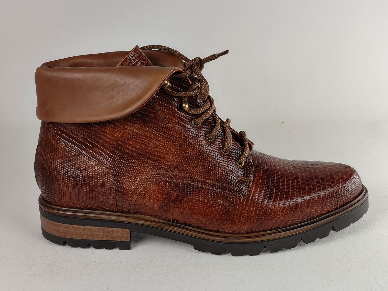 A brown leather lace-up ankle boot with a textured reptile-patterned side panel, a smooth brown collar, and a black lug sole, shown against a light gray background.