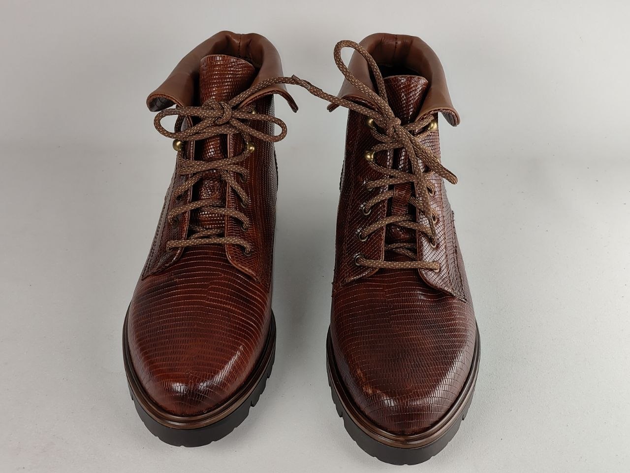 A pair of dark brown textured leather lace-up boots with a glossy finish, photographed against a neutral light gray background.