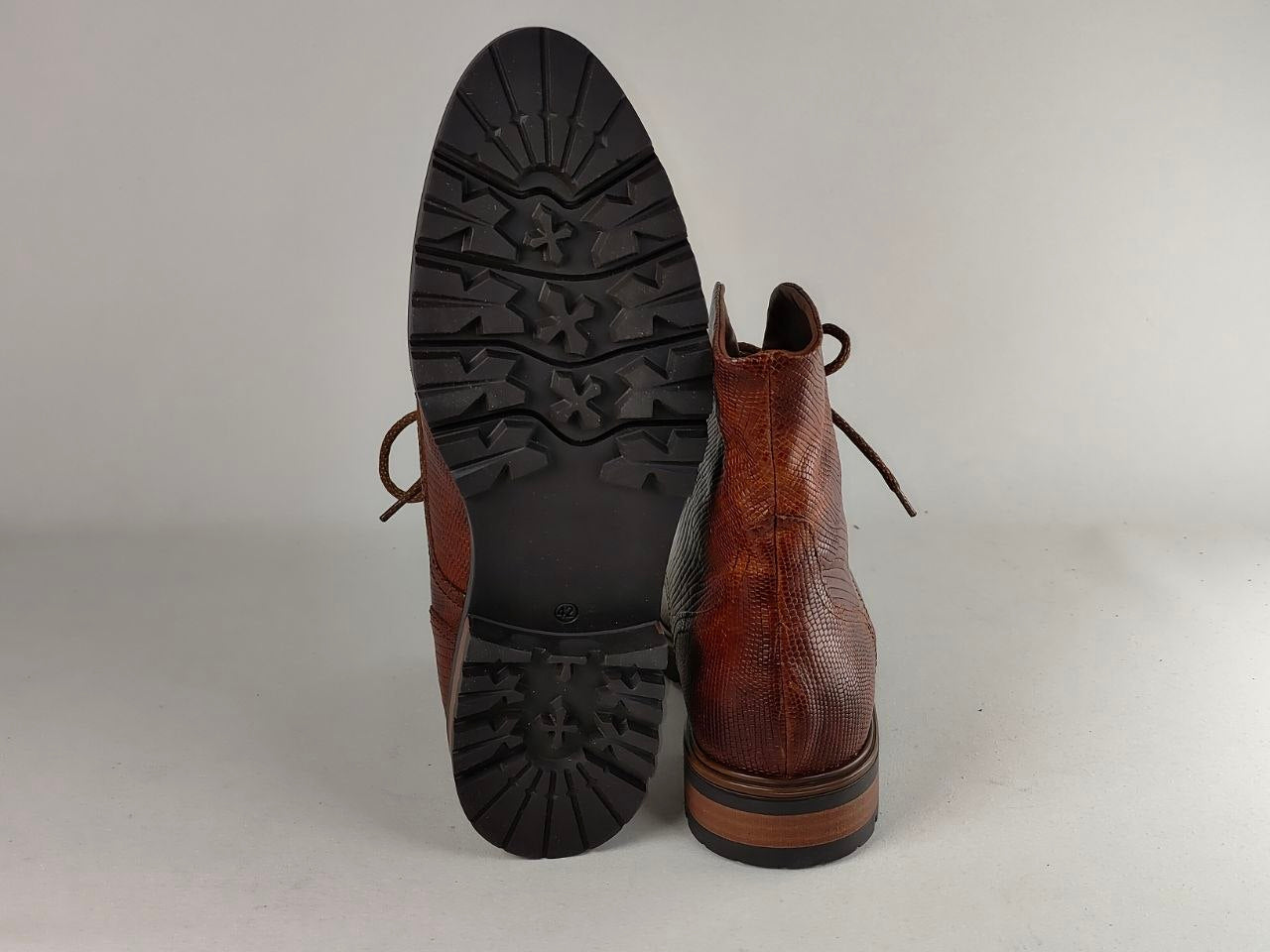A pair of brown leather lace-up boots shown from the back, with one boot standing upright to display the textured brown leather upper and the other lying on its side to show the black treaded outsole.
