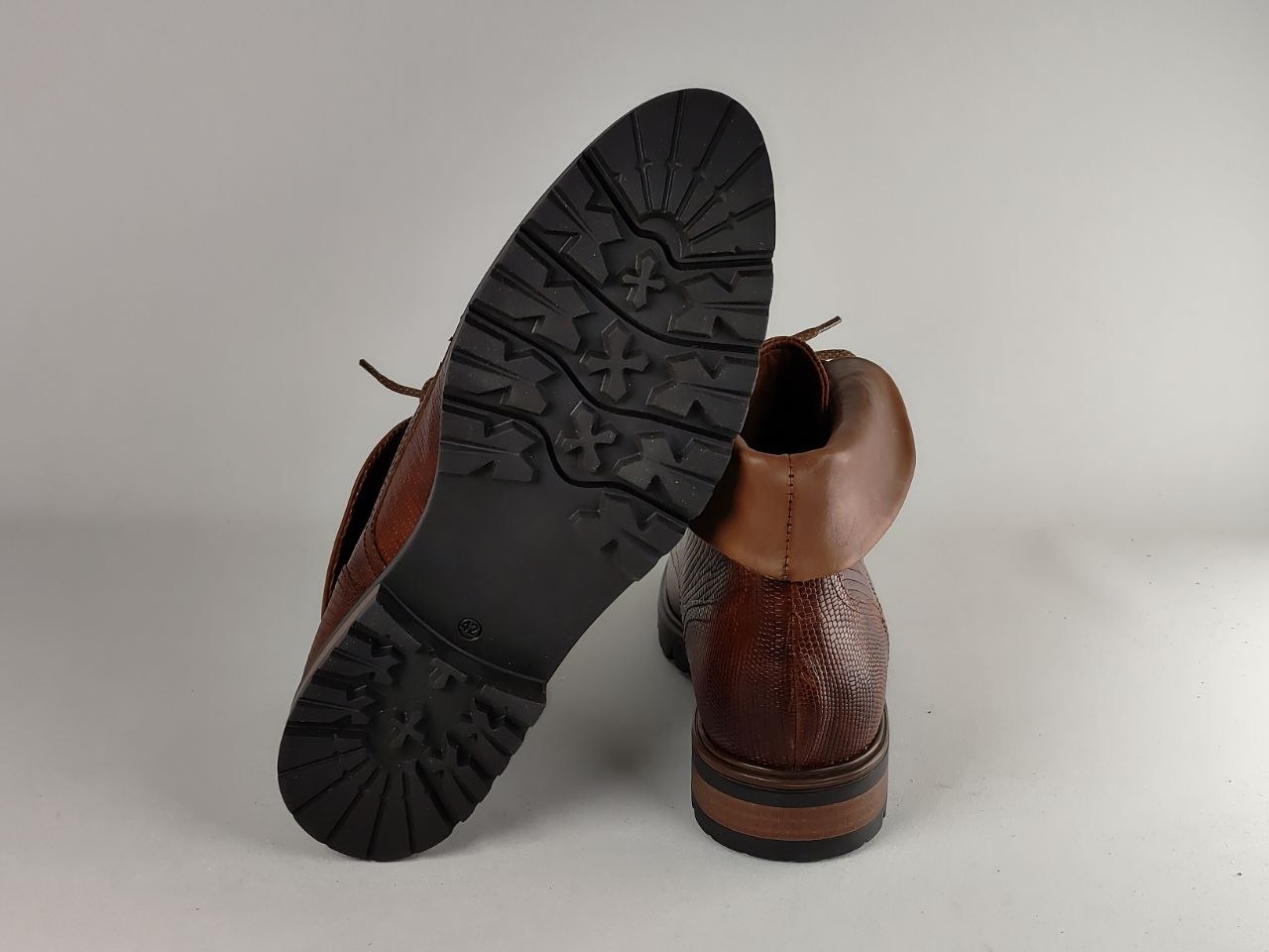 A pair of brown leather ankle boots shown on a light gray studio background, with one boot standing upright and the other tilted to display the black treaded outsole.