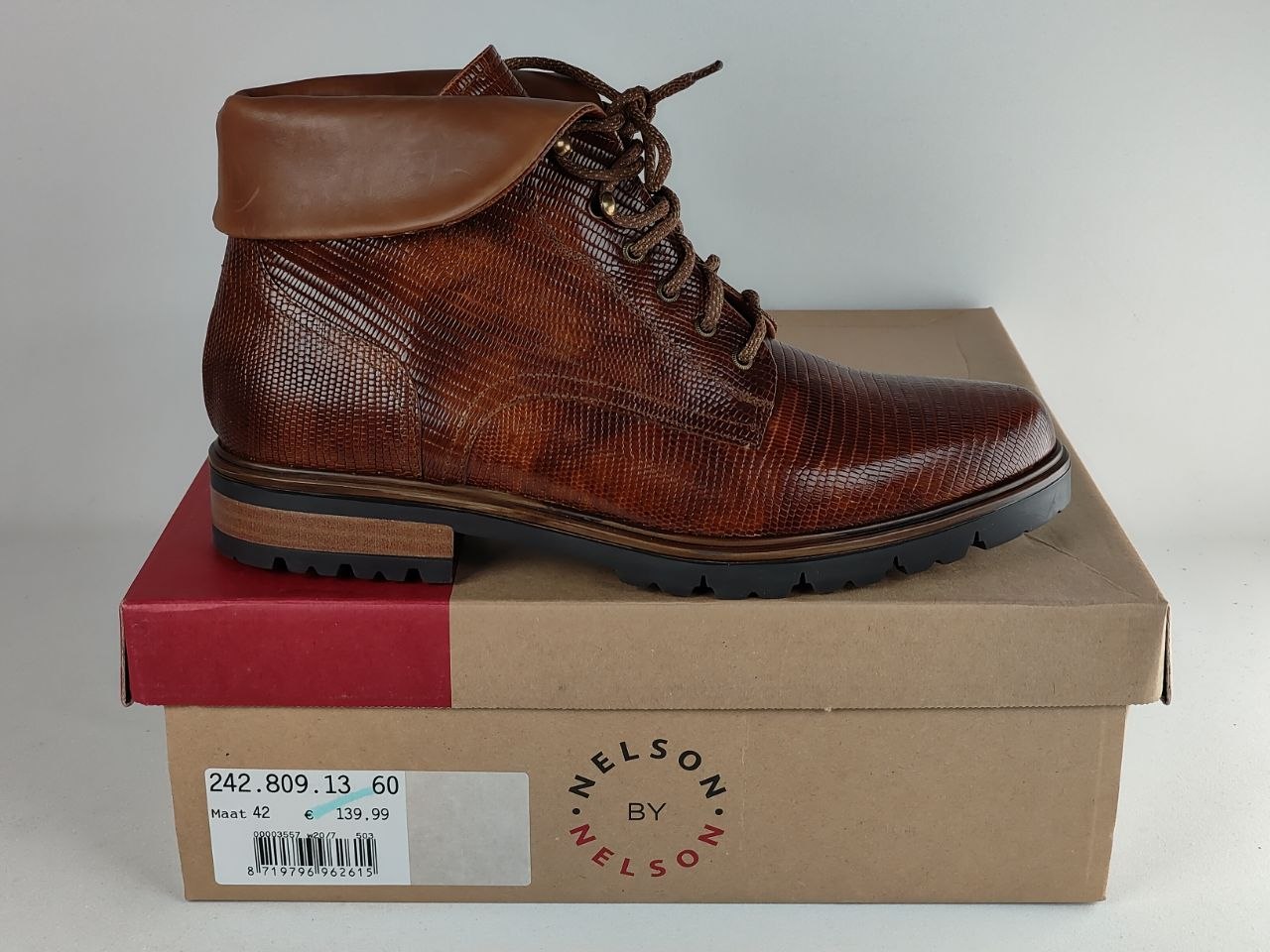 A brown textured leather lace-up ankle boot with a pull tab, resting on top of its cardboard Nelson by Nelson shoebox against a light gray background.