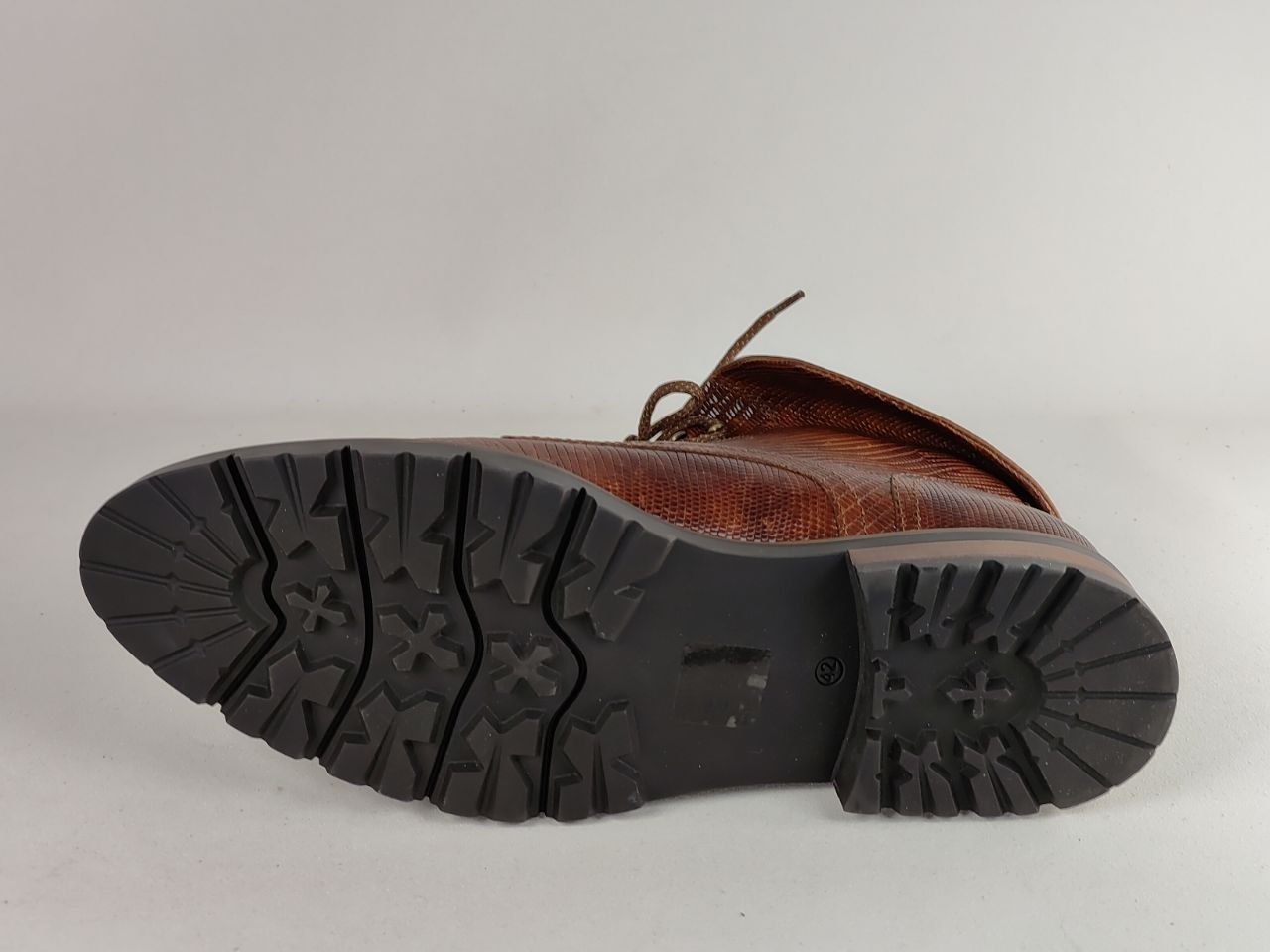 A close-up shot of the treaded black outsole of a brown leather lace-up boot, showing the lug pattern and heel detail against a light gray background.