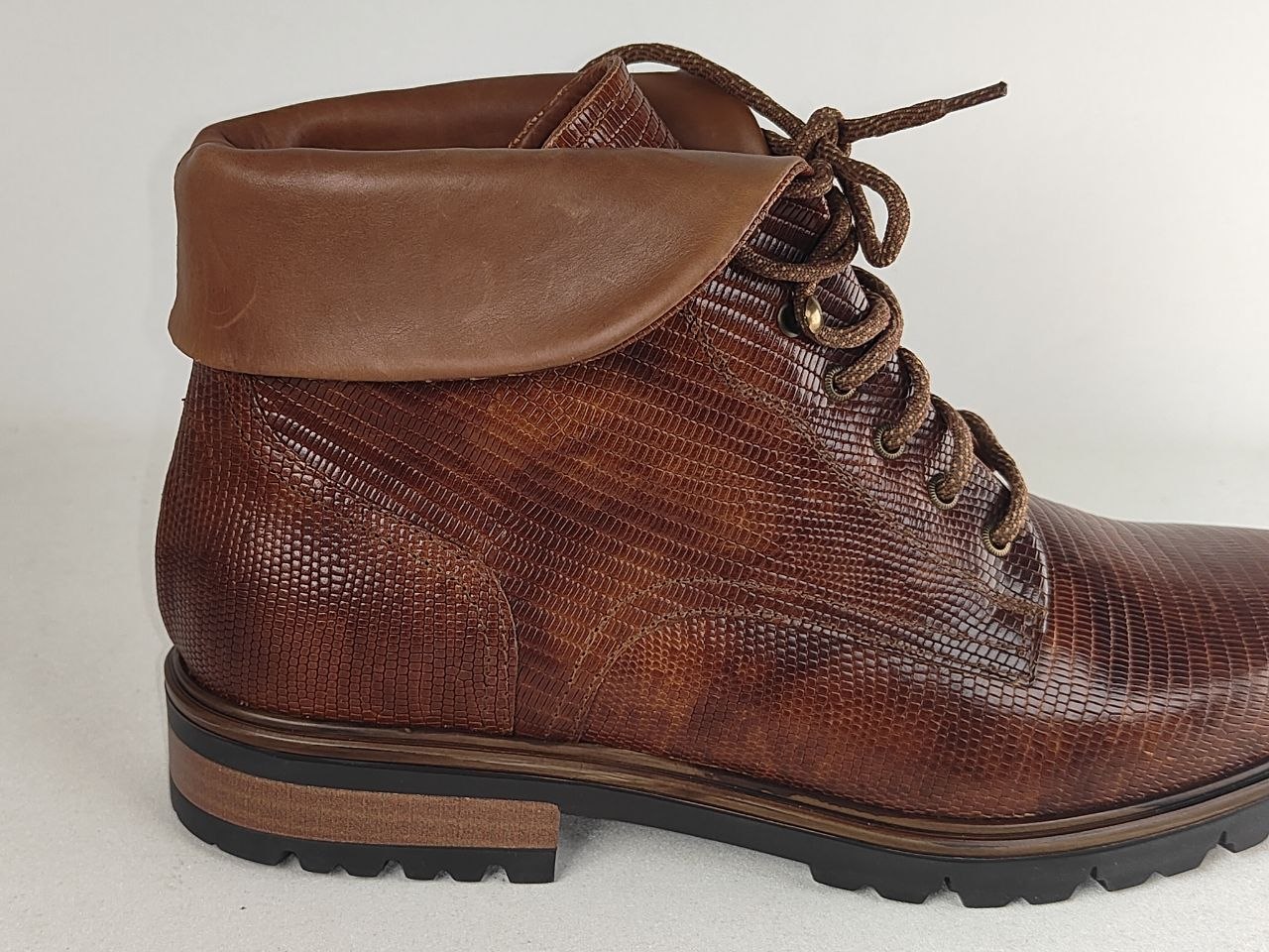 A brown leather lace-up ankle boot with a textured reptile-patterned side panel, a smooth brown collar, and a black lug sole, shown against a plain light background.