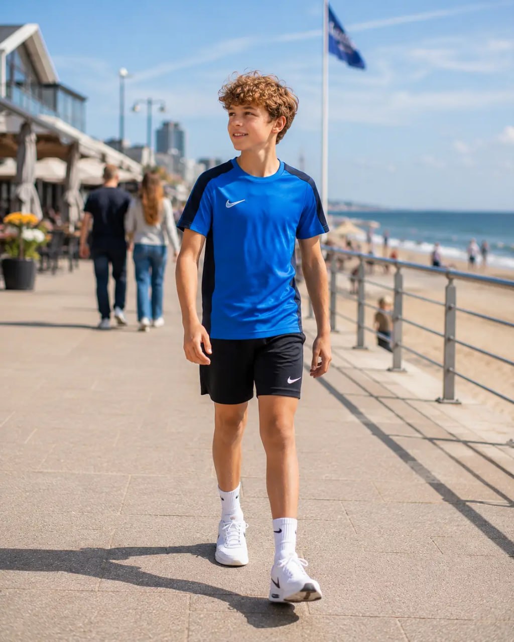 Young person wearing a blue Nike shirt and black shorts walking on a boardwalk by the beach.