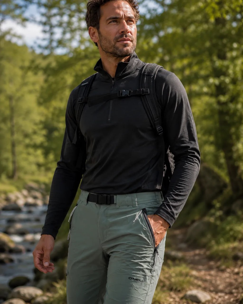A man in black jachet and a green ONeill Hybrid Soft shell Outdoor Pants standing at the riverside with a back ground of jungle.