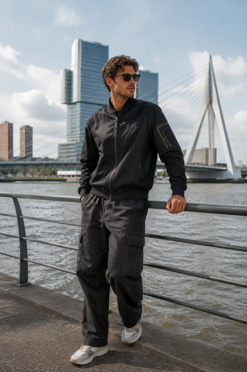 Man in black outfit standing by a waterfront with modern buildings in the background