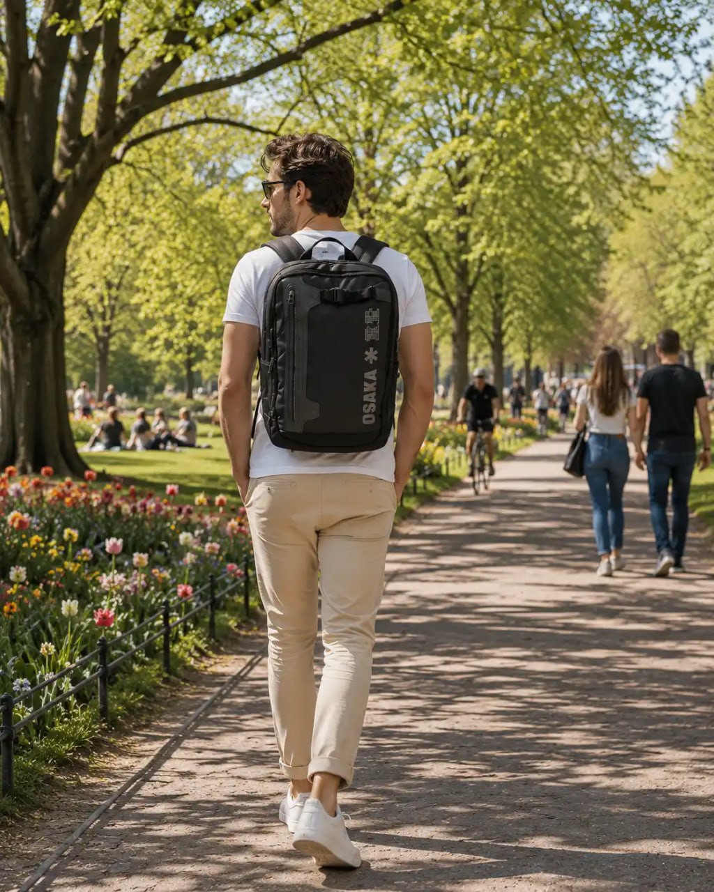 Man walking in a park with a black backpack, surrounded by trees and people.