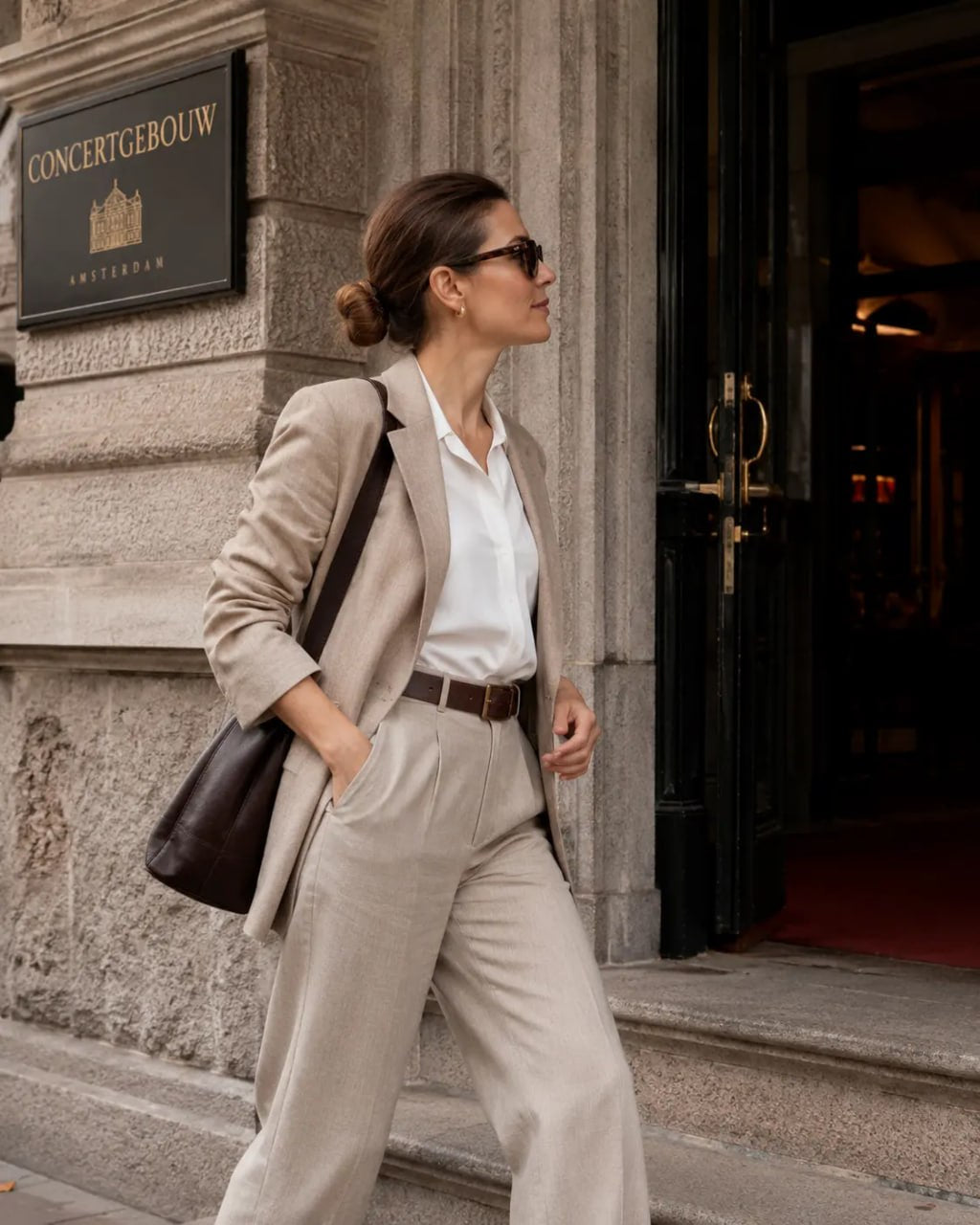 Woman in a beige suit standing outside a building with a sign that reads 'Concertgebouw'.