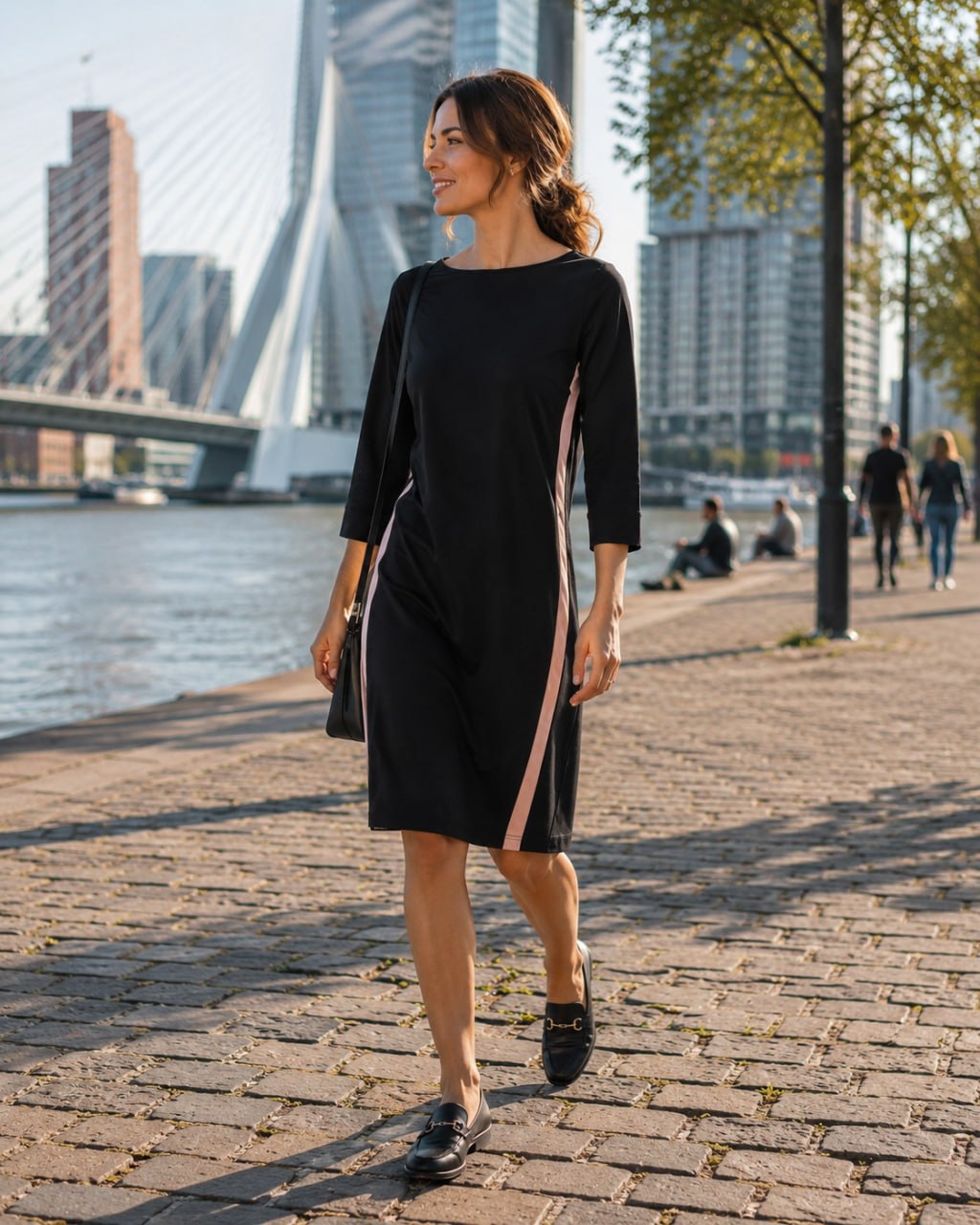 Woman in a black dress standing on a brick path by a waterfront with modern buildings in the background.