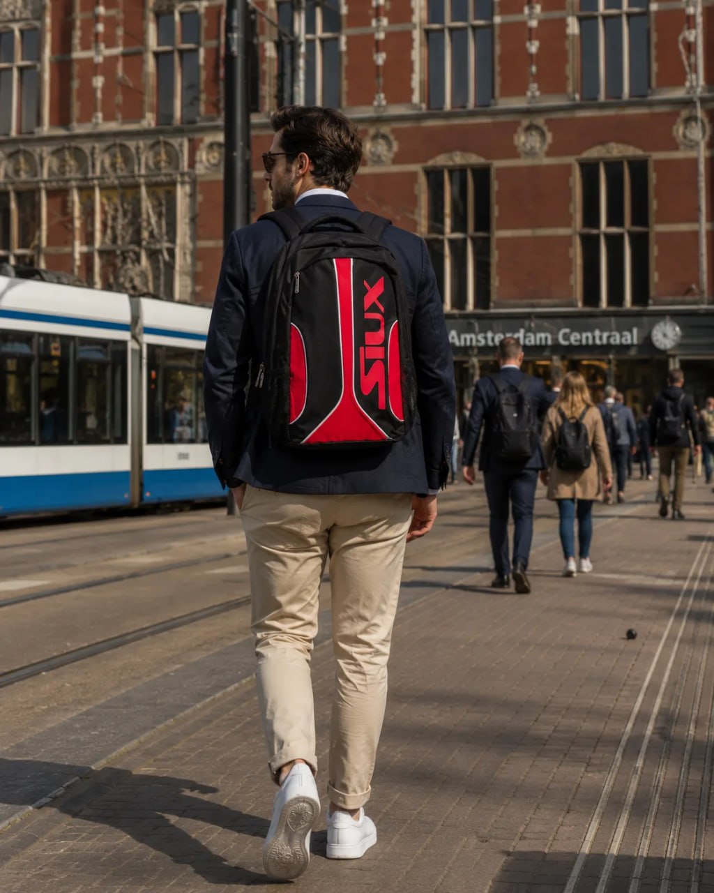 Man walking on a city street with a red and black backpack, Amsterdam Central station in the background.