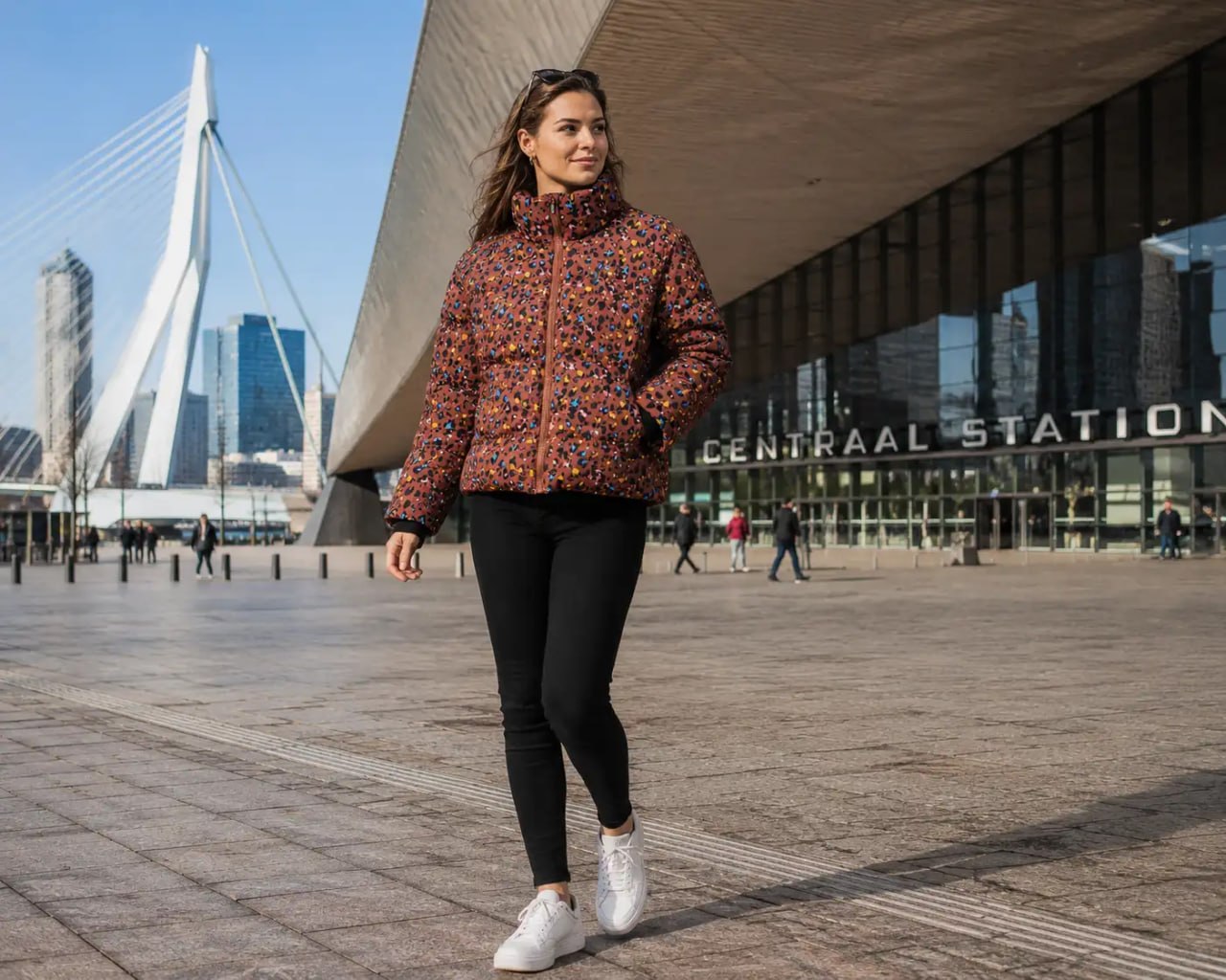 Woman in a colorful patterned Stronger heat short jacket standing in front of Centraal Station, with a modern architectural structure and city skyline in the background.