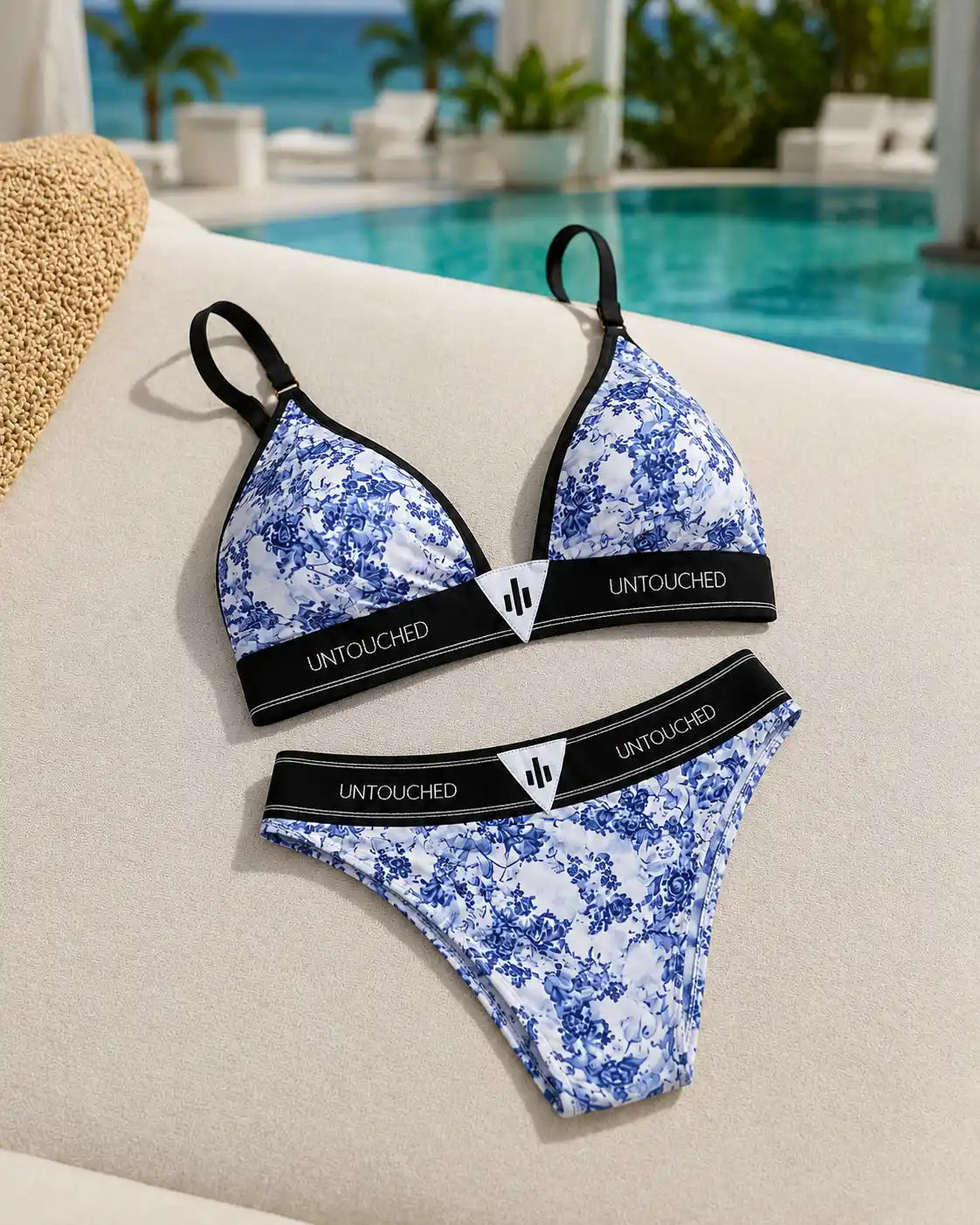 Blue and white floral print bikini set with black trim and 'UNTOUCHED' branding on the waistbands, displayed on a beige lounge chair next to a pool with a beach background.