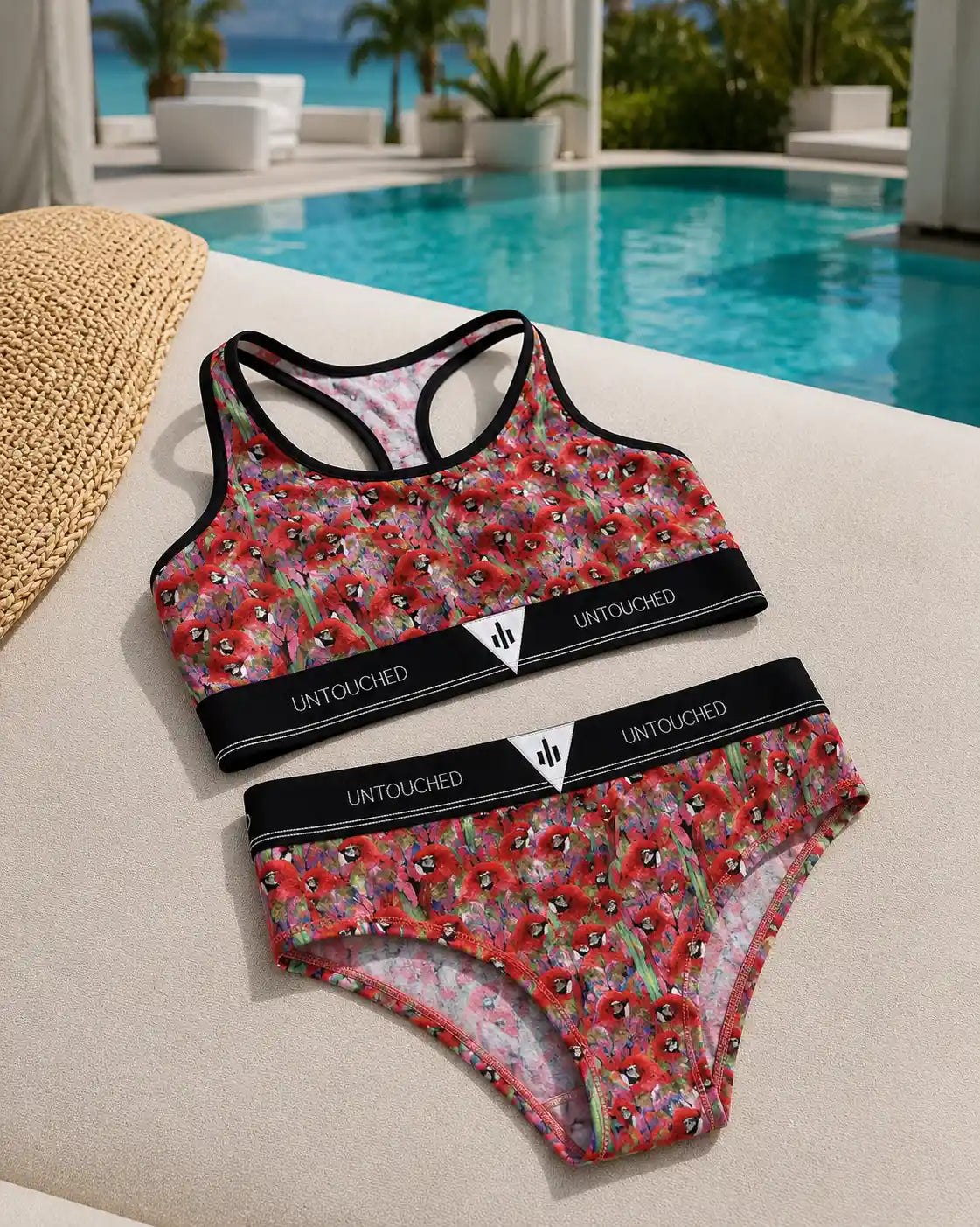 A two-piece bikini set laid flat on a beige lounge chair beside a swimming pool. The set features a vibrant red, pink, and green floral pattern with black trim and a black waistband printed with the brand name 'UNTOUCHED'.