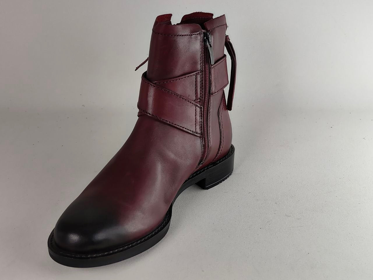 A single dark burgundy leather ankle boot with a black sole, featuring a side zipper and decorative strap details, photographed against a neutral light gray background.