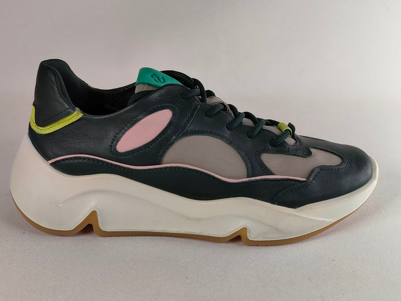 A low-top sneaker with a black leather and gray mesh upper, featuring a chunky white midsole, a light pink accent line, a small pink oval on the side, and a bright lime green heel accent.